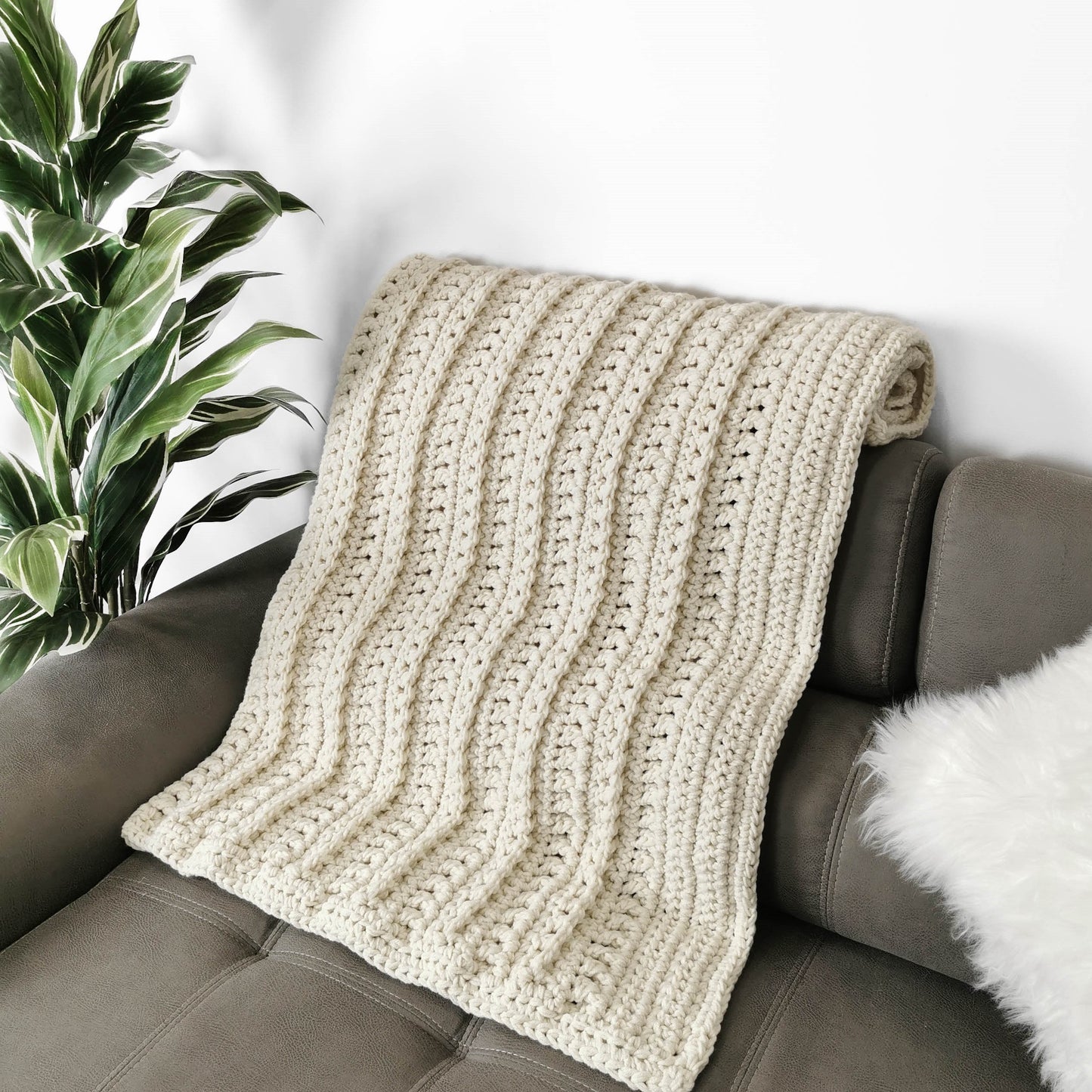 May include: A cream-colored crocheted throw blanket draped over a brown couch. The blanket has a textured, ribbed pattern.