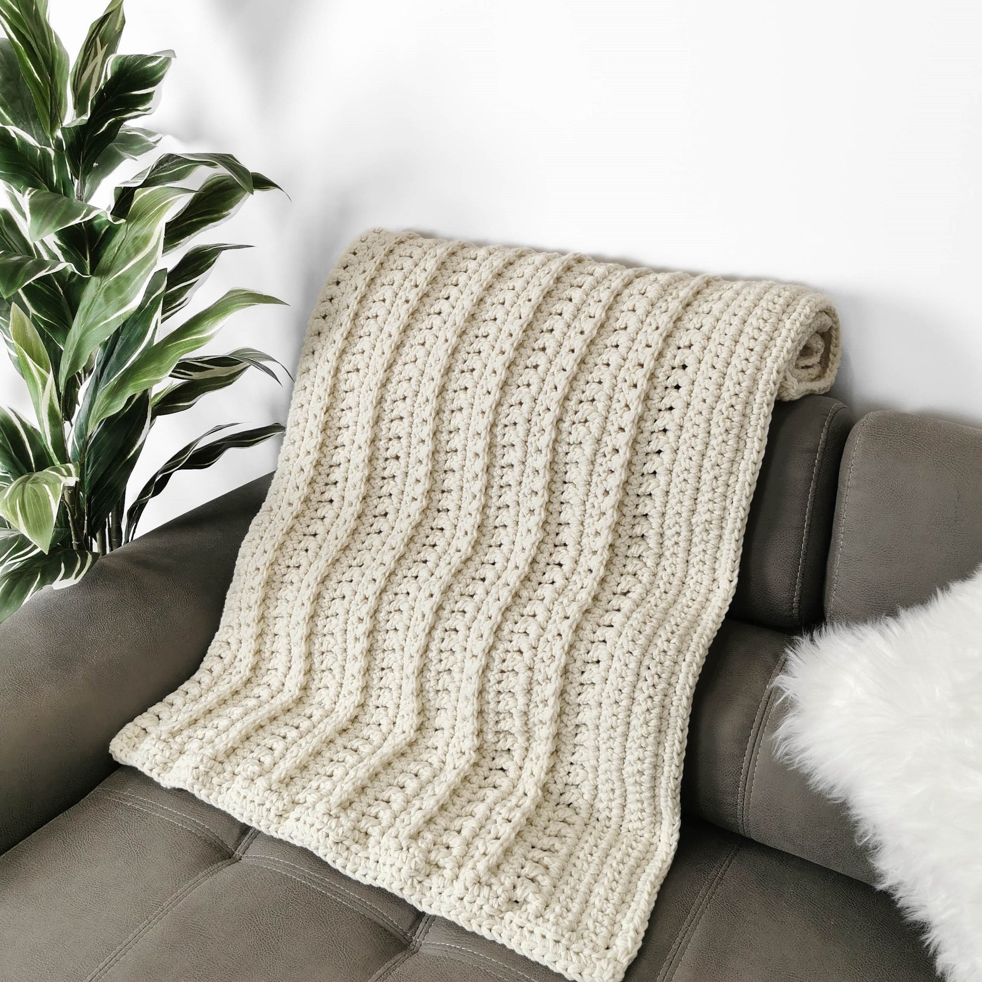 May include: A cream-colored crocheted throw blanket draped over a brown couch. The blanket has a textured, ribbed pattern.