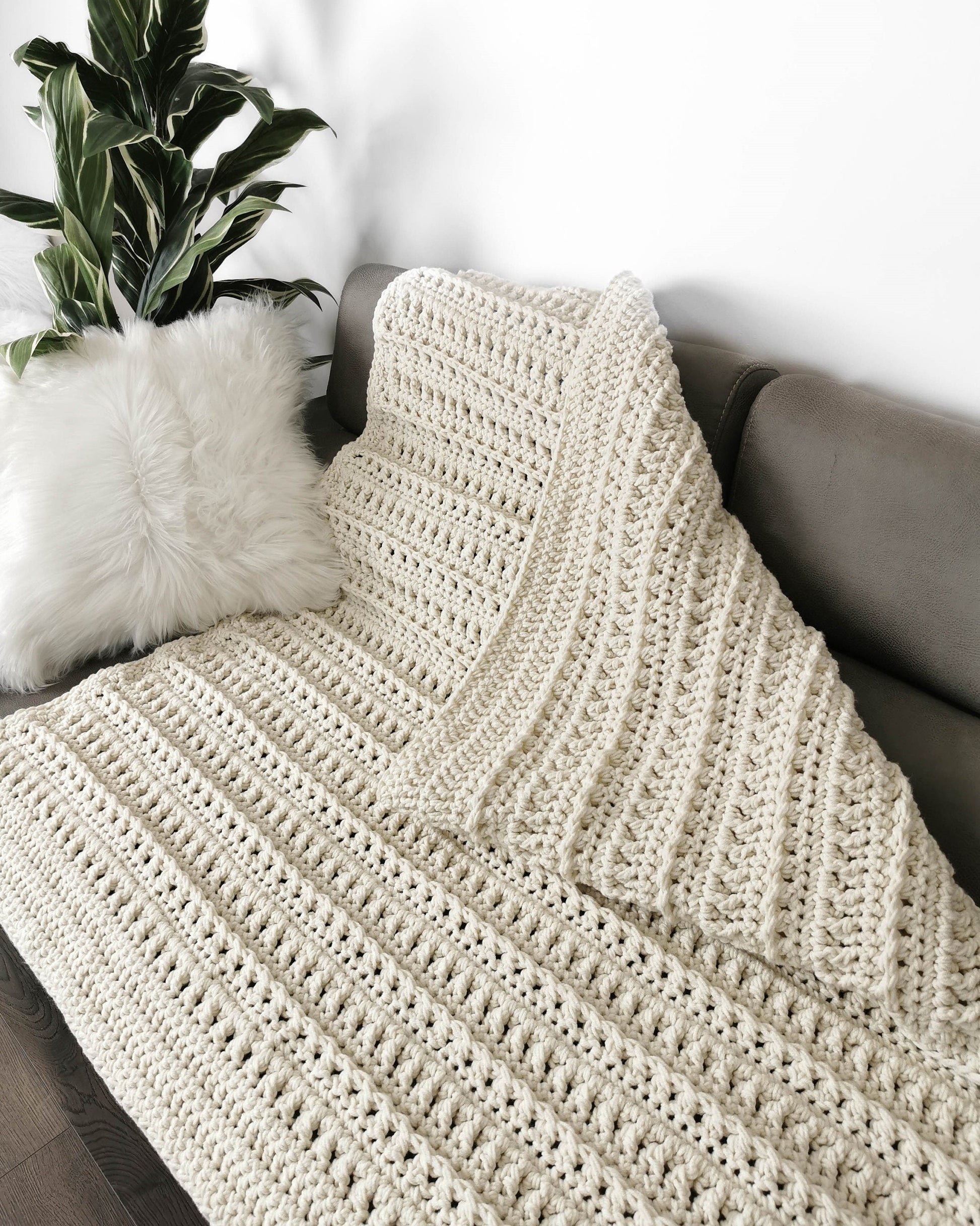May include: A cream-colored crocheted throw blanket draped over a couch with a white faux fur pillow. The blanket has a textured stitch pattern.