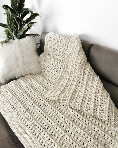 May include: A cream-colored crocheted throw blanket draped over a couch with a white faux fur pillow. The blanket has a textured stitch pattern.
