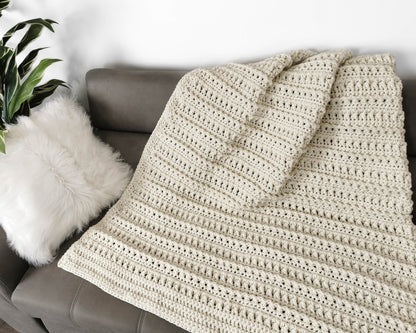 May include: A cream-colored crocheted throw blanket draped over a gray couch. The blanket has a textured, ribbed pattern.