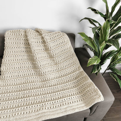 May include: A cream-colored crocheted throw blanket draped over a gray couch. The blanket has a textured stitch pattern and is made with a soft yarn.