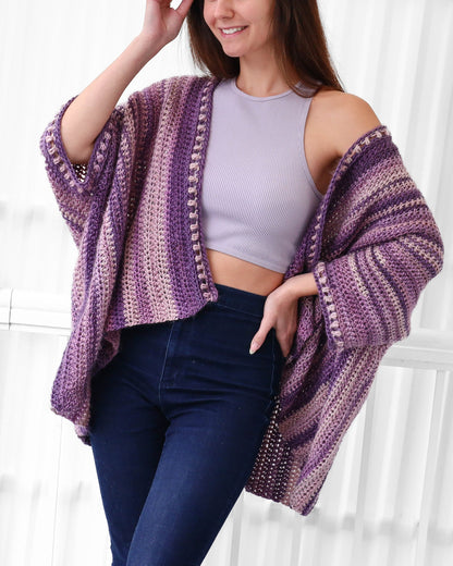 May include: A purple and pink striped crochet cardigan with a crocheted border. The cardigan is open in the front and has long sleeves. The model is wearing the cardigan over a purple tank top and blue jeans.