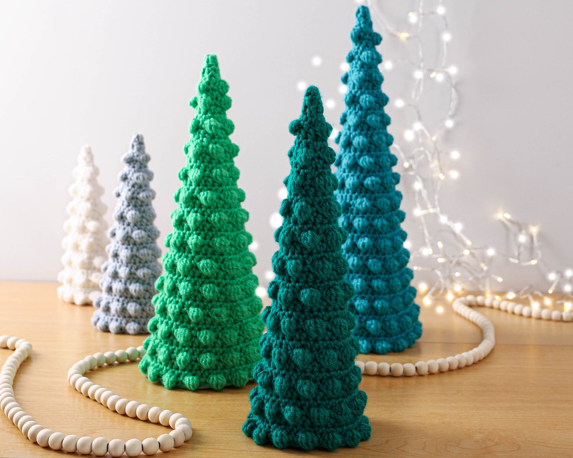 May include: Five crocheted Christmas trees in different sizes and colors. The trees are white, gray, green, and teal. The trees are arranged in a row on a wooden surface. There is a white beaded garland in the foreground.