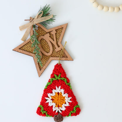 May include: A crocheted Christmas tree ornament in red, white, green, and orange with a brown felt base. The ornament is hanging from a wooden star with the word "JOY" written on it.