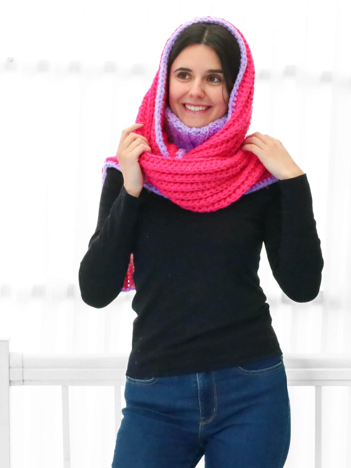 May include: A pink and purple crocheted cowl with a hood. The cowl is worn by a person with dark hair and a black shirt.