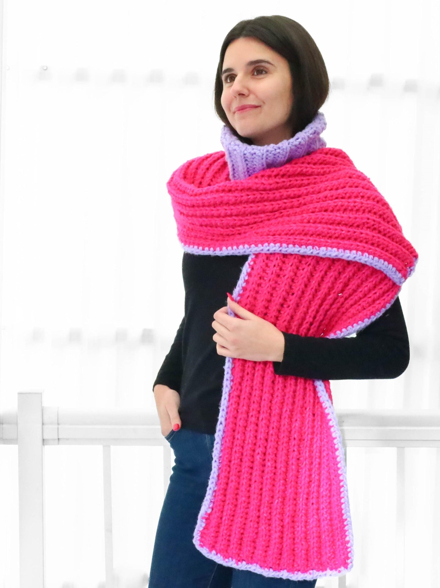 May include: A pink and purple crocheted scarf with a ribbed texture. The scarf is folded and draped around the neck of a person wearing a black turtleneck shirt and blue jeans.