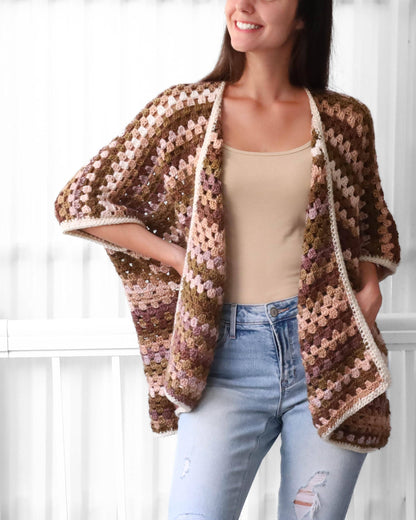 May include: A crocheted cardigan with a brown, pink, and white color scheme. The cardigan has a loose fit and a wide, open neckline. It is made with a granny square stitch pattern.