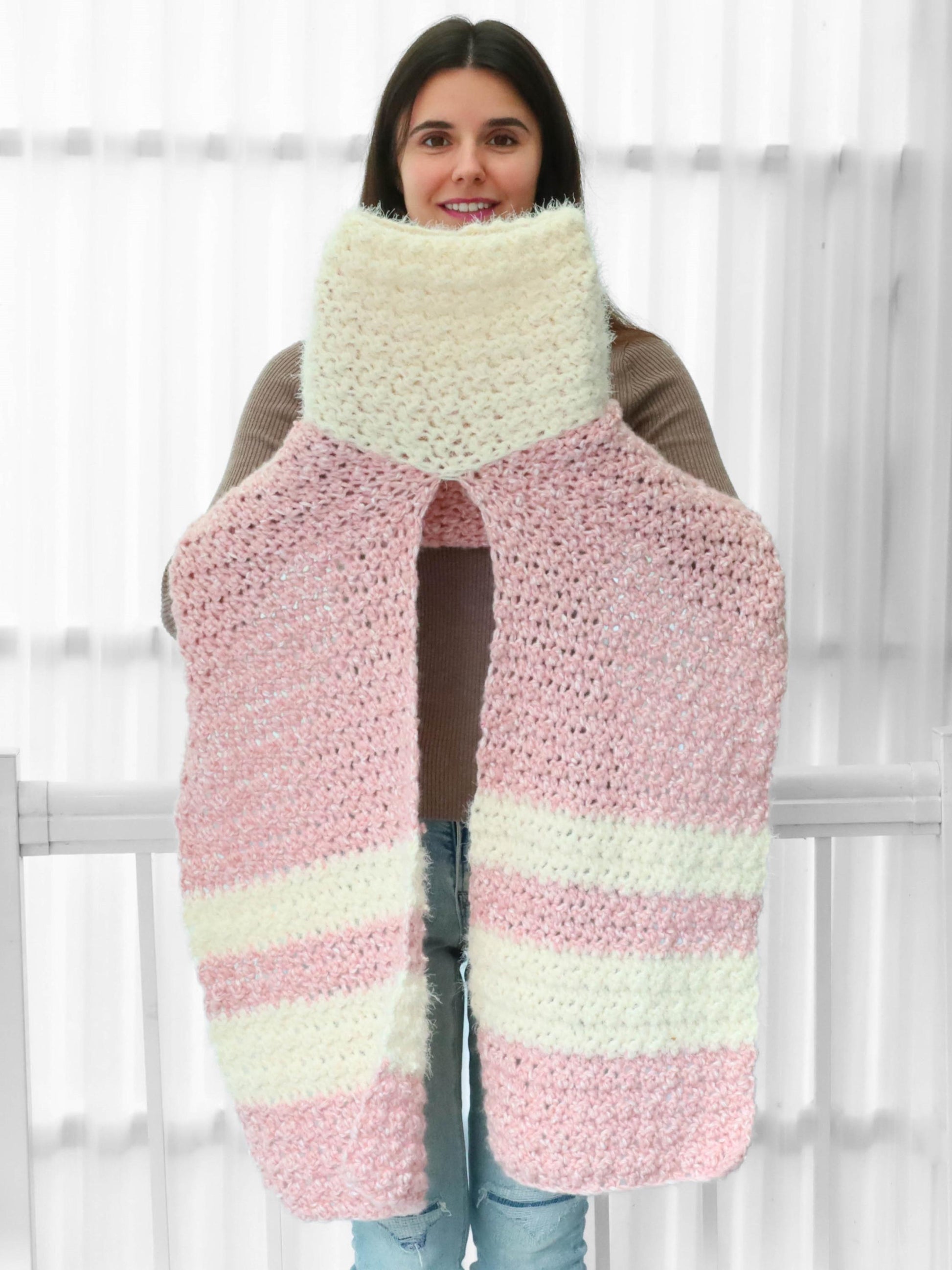 May include: A pink and white striped crocheted scarf with a fluffy texture. The scarf is long and wide, and it is held up by a person.