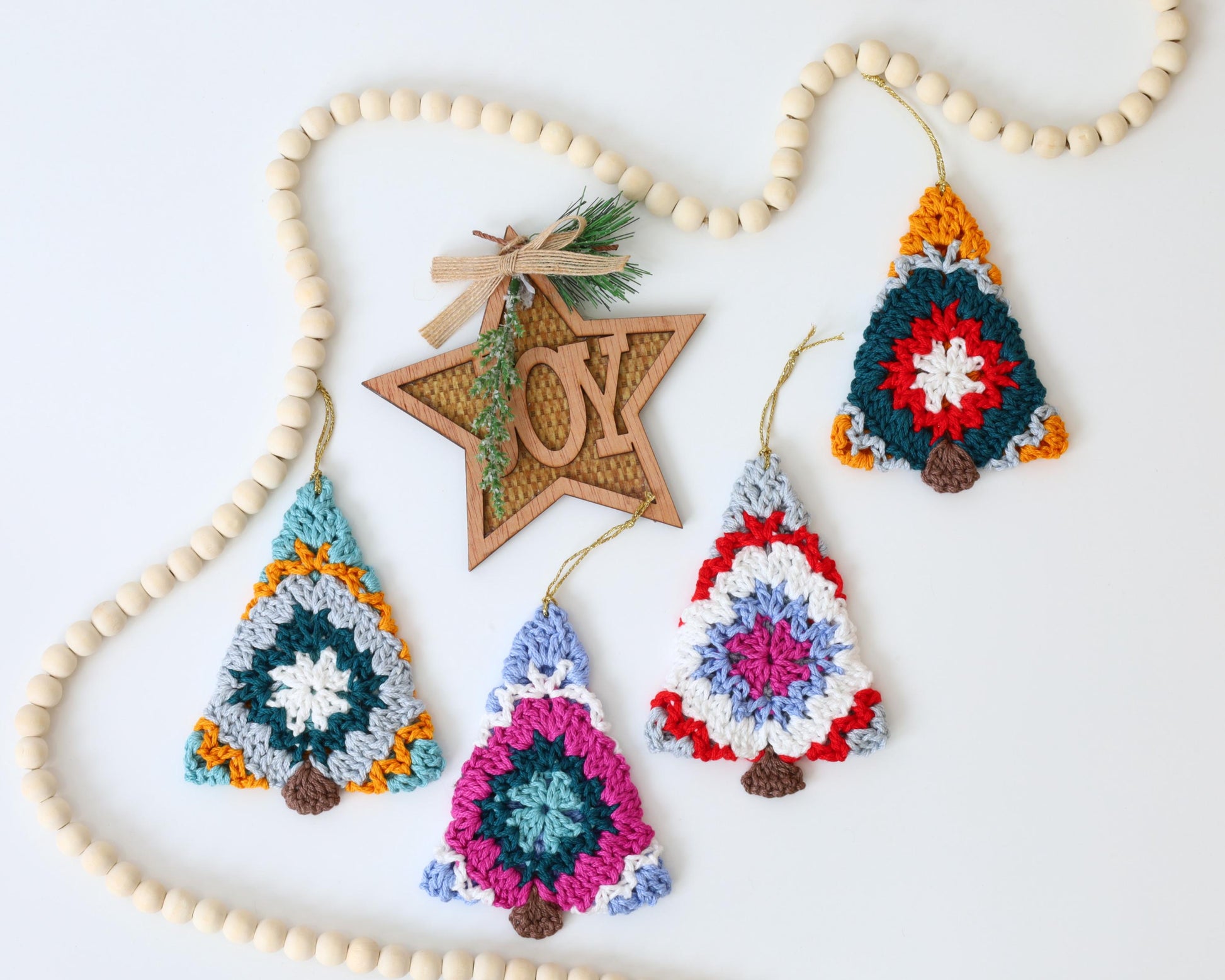 May include: Four crocheted Christmas tree ornaments in various colors, including red, blue, green, yellow, and white. The ornaments are hanging from a wooden bead garland with a wooden star cutout that says "JOY".