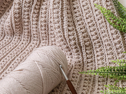 May include: A beige crocheted blanket featuring a textured pattern of vertical ribbed lines and small raised bobble stitches, accompanied by a matching beige yarn ball and a metal crochet hook with a brown handle resting on the fabric.