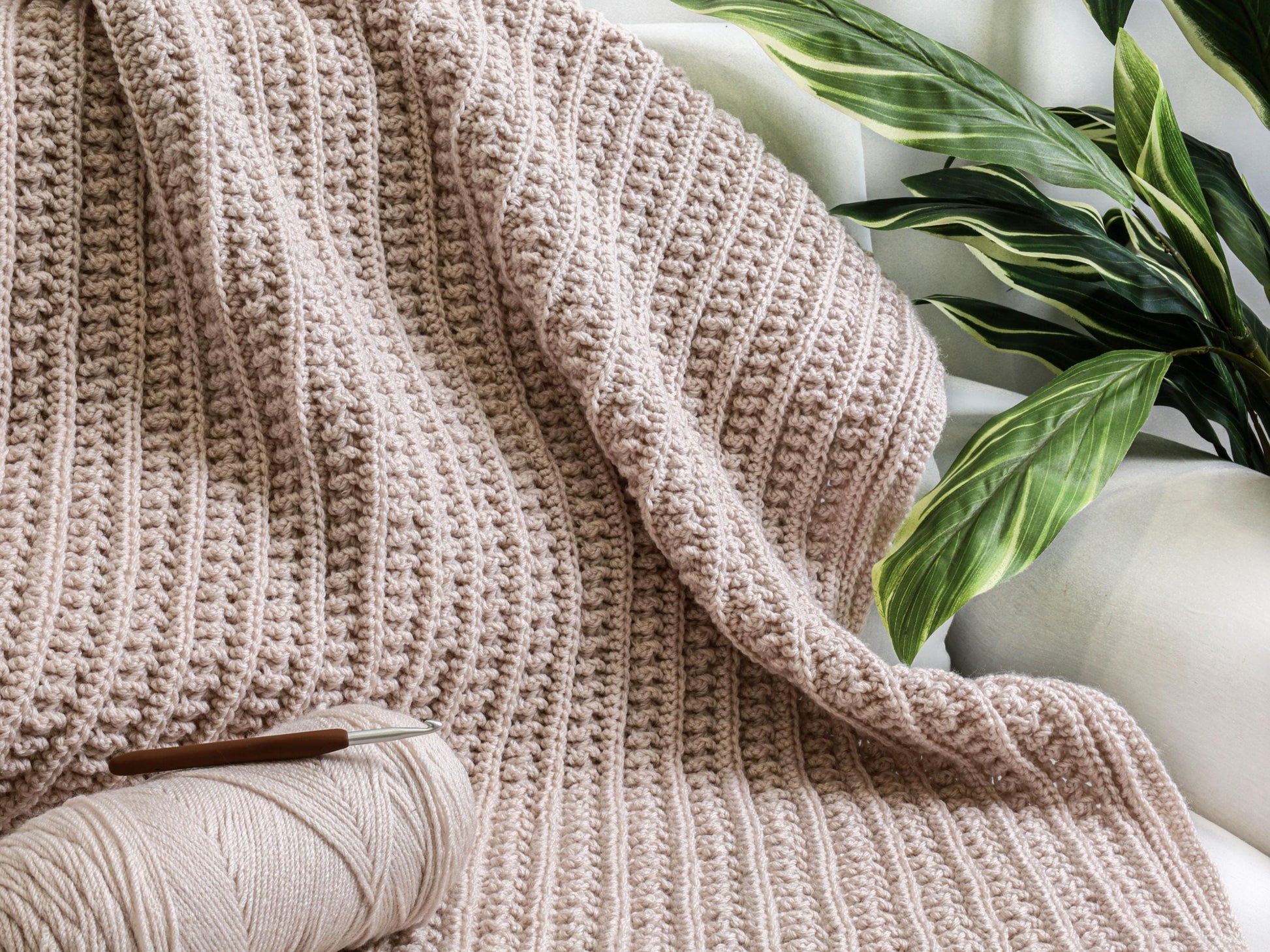 May include: A light pink crocheted blanket featuring a textured ribbed pattern. A matching ball of yarn and a brown-handled crochet hook are placed on the surface of the blanket. The fabric displays a consistent, repeating stitch design throughout.