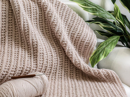 May include: A light pink crocheted blanket featuring a textured ribbed pattern. A matching ball of yarn and a brown-handled crochet hook are placed on the surface of the blanket. The fabric displays a consistent, repeating stitch design throughout.