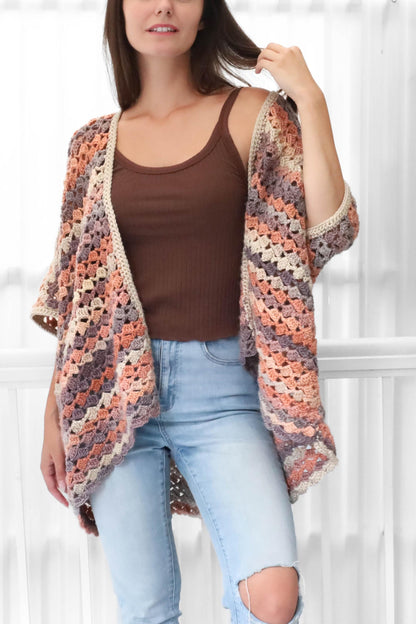 May include: A handmade, open-front crochet cardigan in a granny square pattern. The color palette includes peach, lavender, cream, and brown tones. The sleeves are elbow-length. The cardigan is paired with a brown tank top and light blue jeans.