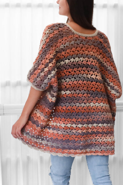 May include: A multi-colored crochet poncho in shades of orange, purple, and gray. The open weave garment has a round neckline and short sleeves. The poncho is worn over blue jeans.