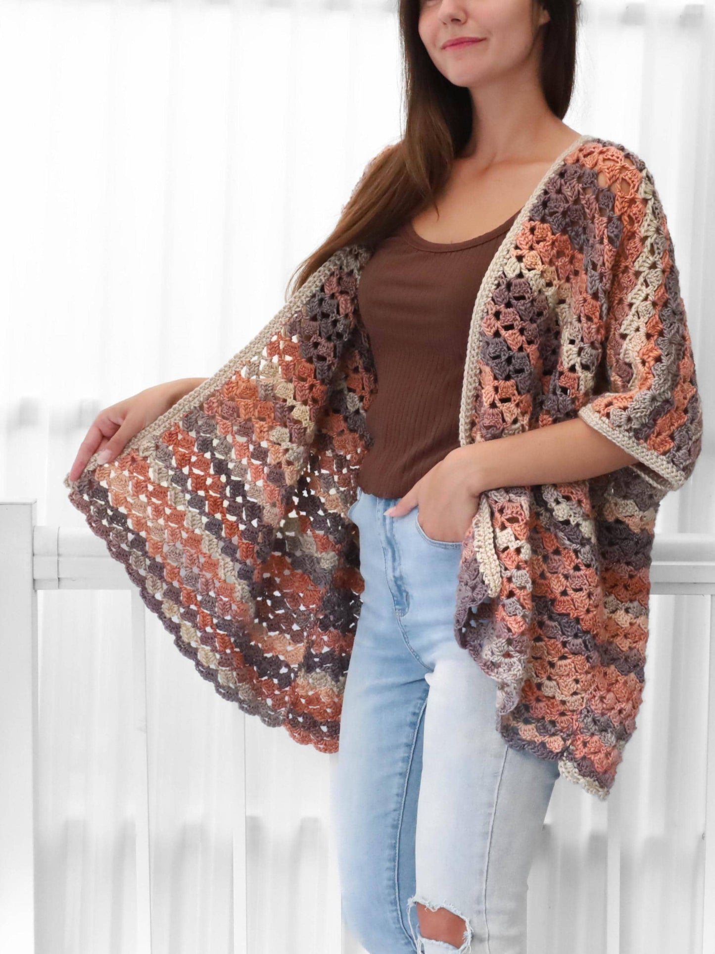 May include: A multi-colored crochet shawl in shades of peach, gray, and beige. The open-front design features a scalloped edge and is worn over a brown top and blue jeans. The shawl is a handmade garment.