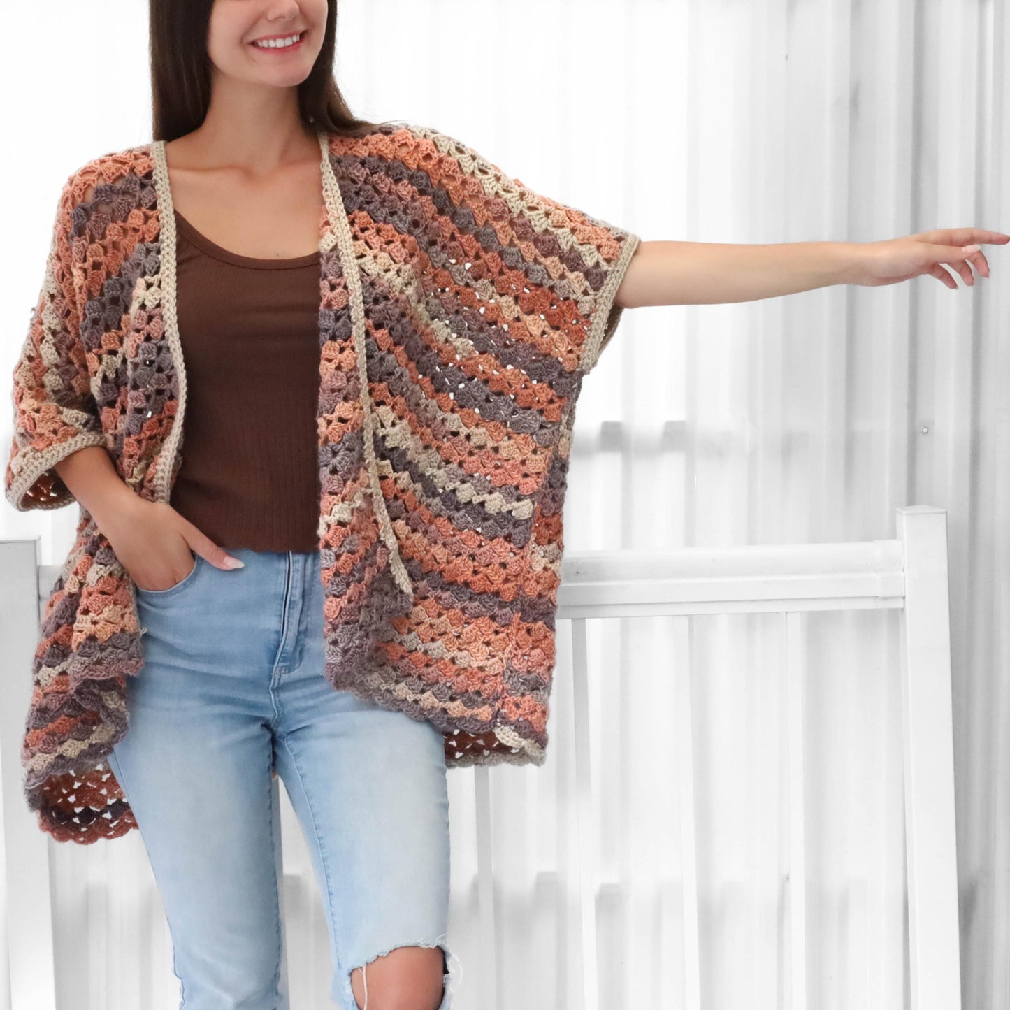 May include: A multi-colored crochet cardigan in shades of peach, gray, and brown. The open-front cardigan has short sleeves and a beige trim. The woman is wearing a brown top and light blue jeans.