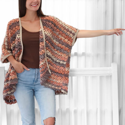 May include: A multi-colored crochet cardigan in shades of peach, gray, and brown. The open-front cardigan has short sleeves and a beige trim. The woman is wearing a brown top and light blue jeans.