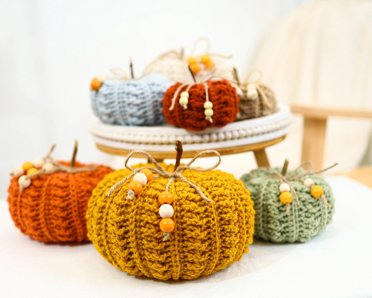May include: A collection of handmade crochet pumpkins in various colors, including orange, yellow, and green. Each pumpkin features a stem and is adorned with wooden beads. Some pumpkins are displayed on a decorative stand, creating a fall-themed display.