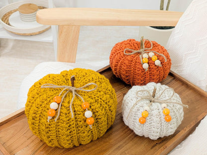 May include: Three crocheted pumpkins in shades of yellow, orange, and white, arranged on a wooden tray. Each pumpkin has a twine stem and is adorned with wooden beads. The pumpkins are a decorative item for the fall season.