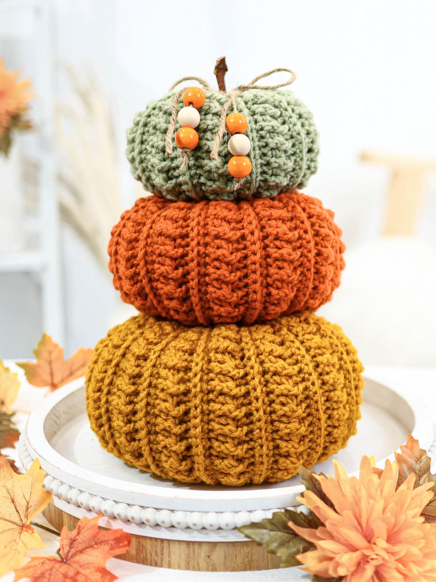 May include: A stack of three crocheted pumpkins in shades of green, orange, and yellow. The top pumpkin is green with wooden beads and a twine stem. The middle pumpkin is orange, and the bottom pumpkin is yellow. The pumpkins are stacked on a white tray.