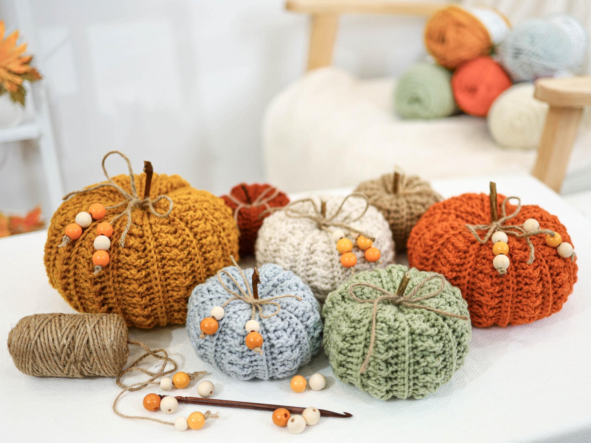May include: A collection of handmade crochet pumpkins in various colors, including orange, yellow, white, and green. Each pumpkin has a twine stem and is adorned with wooden beads. A ball of twine and a crochet hook are also present.
