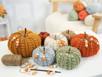 May include: A collection of handmade crochet pumpkins in various colors, including orange, yellow, white, and green. Each pumpkin has a twine stem and is adorned with wooden beads. A ball of twine and a crochet hook are also present.