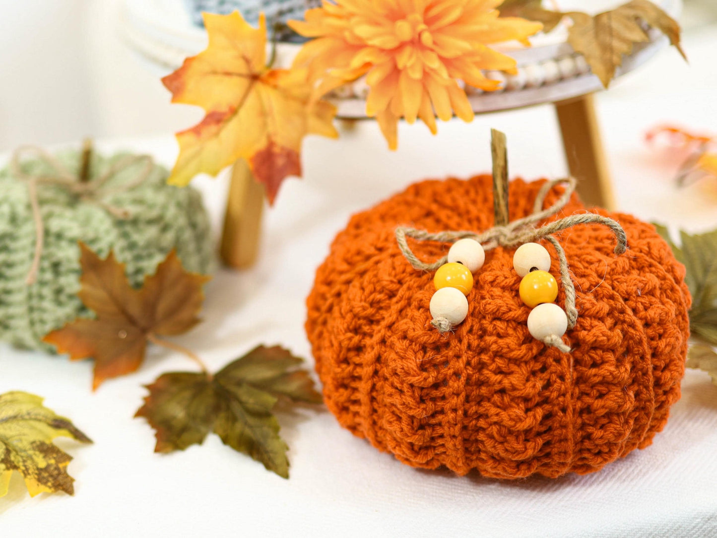May include: An orange crocheted pumpkin with a twine stem and wooden beads. The pumpkin is surrounded by fall foliage, including leaves and a flower. A green crocheted pumpkin is in the background.