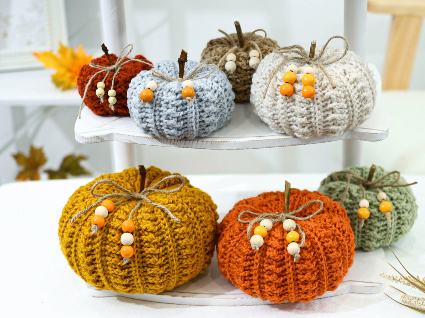 May include: A collection of handmade crochet pumpkins in various colors, including orange, yellow, and green. Each pumpkin has a twine stem, a twine bow, and decorative wooden beads. These fall-themed decorations are displayed on a white shelf.