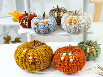 May include: A collection of handmade crochet pumpkins in various colors, including orange, yellow, and green. Each pumpkin has a twine stem, a twine bow, and decorative wooden beads. These fall-themed decorations are displayed on a white shelf.