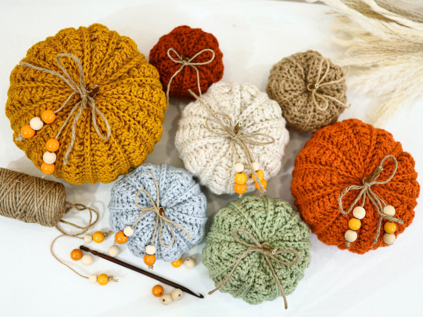 May include: A collection of handmade crochet pumpkins in various colors, including mustard yellow, orange, and light blue. Each pumpkin is adorned with twine and wooden beads, creating a rustic and autumnal aesthetic. Perfect for fall decor.