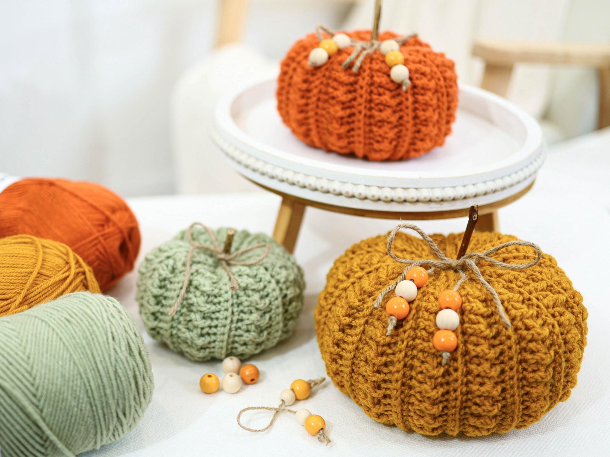 May include: A collection of handmade crochet pumpkins in various colors, including orange, green, and gold. Each pumpkin features a stem and decorative accents like wooden beads. Yarn skeins in coordinating colors are also present, suggesting a crafting theme.