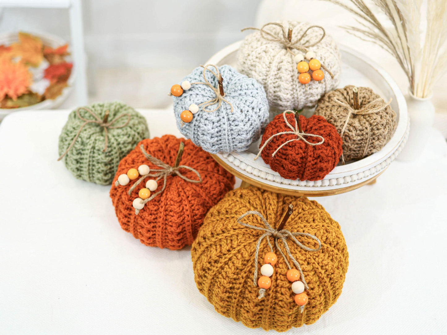 May include: A collection of handmade crochet pumpkins in various colors, including orange, green, and cream. Each pumpkin is adorned with twine and wooden bead accents, creating a rustic and autumnal aesthetic. Some pumpkins are displayed on a white tray.