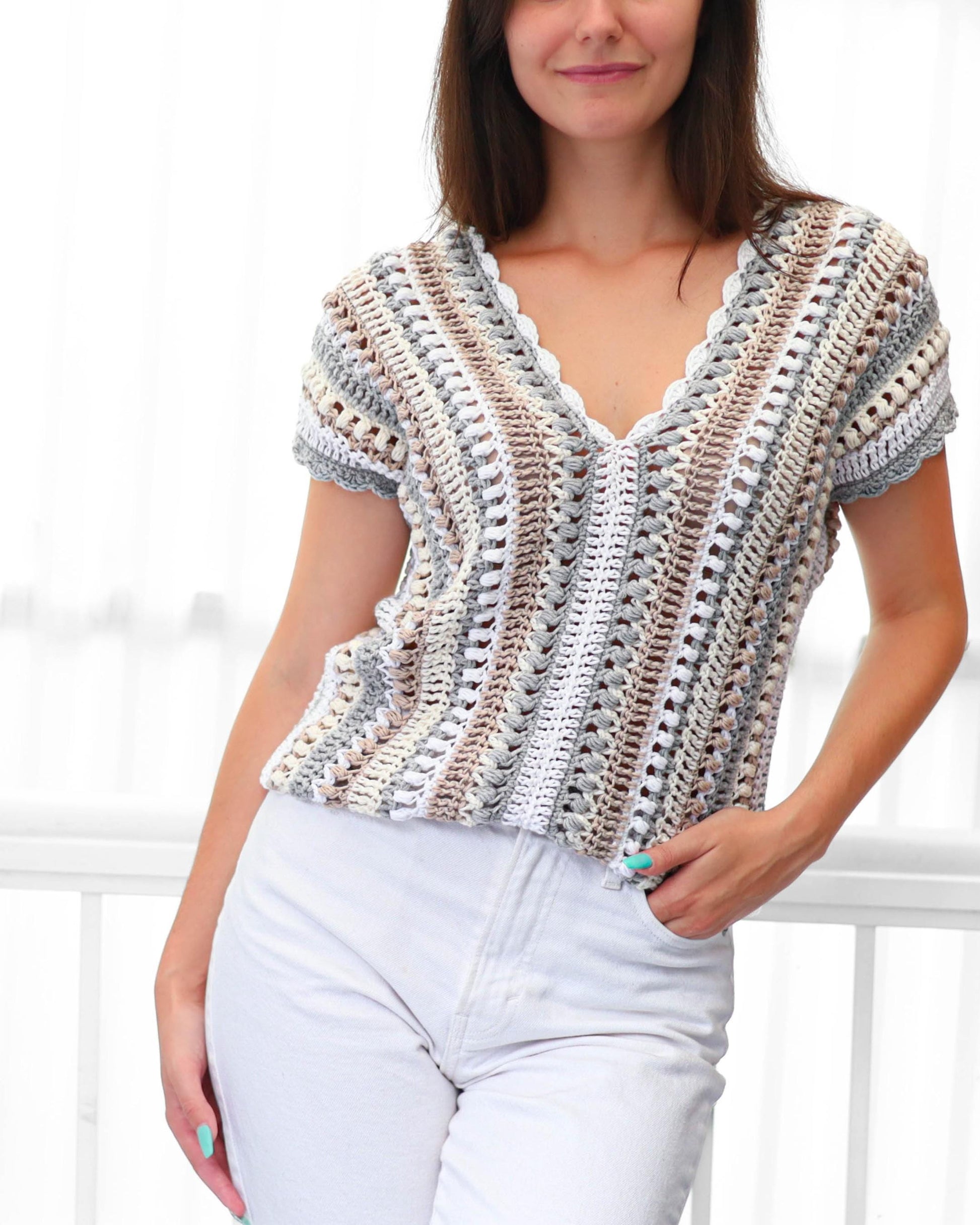 May include: A short-sleeved, V-neck crochet top in a neutral color palette of white, gray, and brown. The top features a scalloped edge along the neckline and sleeves. The crochet pattern creates a textured, open weave design.