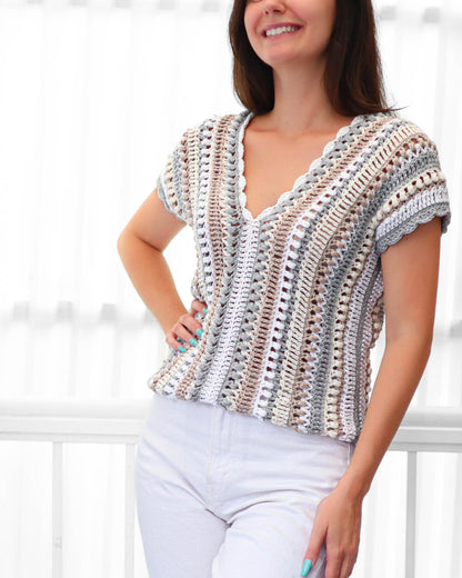 May include: A short-sleeved, crochet top in a neutral color palette of white, gray, and beige. The top features a V-neckline and a scalloped edge. The crochet design has vertical stripes.