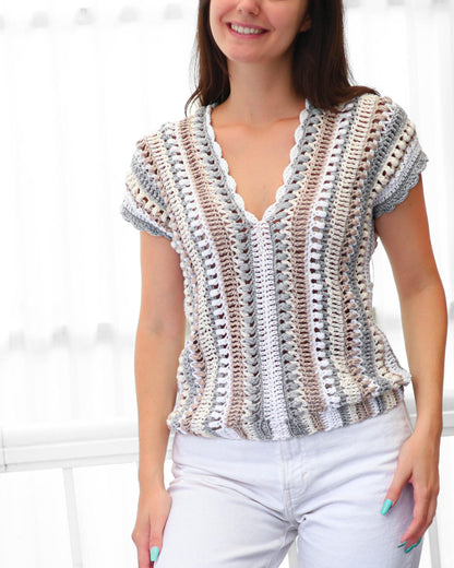 May include: A short-sleeved crochet top in a neutral color palette of white, gray, and brown. The top features a V-neckline with a scalloped edge and a slightly gathered waist. The crochet pattern creates a textured, open design.
