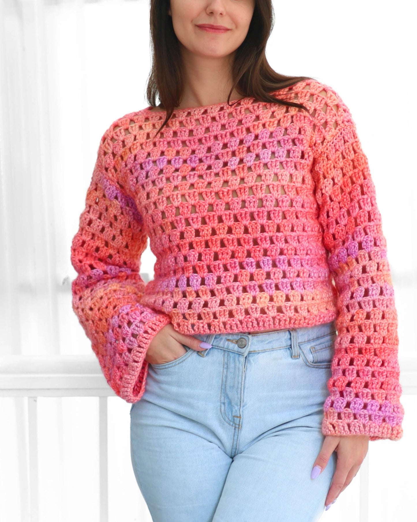 May include: A crochet sweater in shades of pink, orange, and purple. The long-sleeved top has a loose, open weave and a boat neck. The model is wearing light blue jeans. The sweater's color gradient adds a vibrant touch to the overall look.