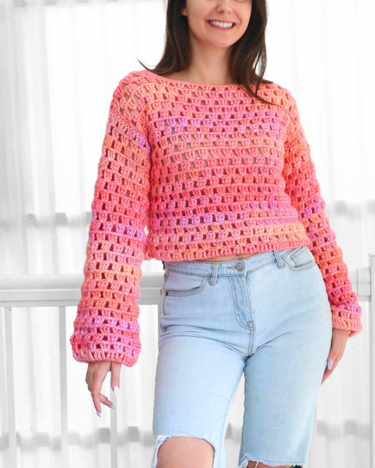 May include: A crochet sweater in shades of pink and orange. The long-sleeved top has a loose, open weave design. The sweater is paired with light blue denim jeans.