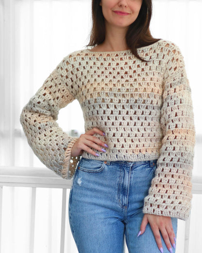 May include: A cream-colored crochet top with long sleeves and a square neckline. The open weave design features a gradient of colors, transitioning from cream to light peach and blue. The top is paired with blue jeans.