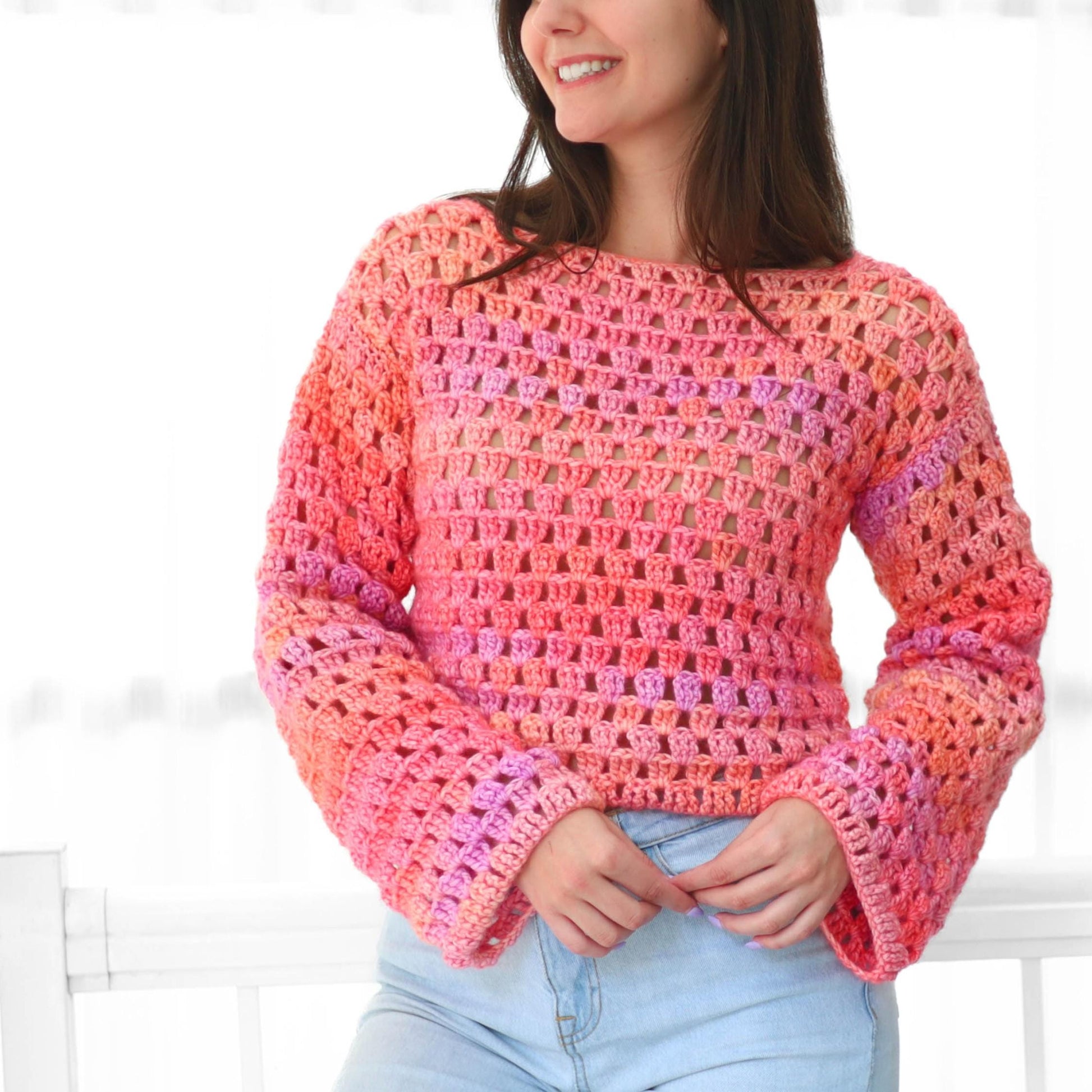 May include: A crocheted sweater in shades of pink, orange, and purple. The long-sleeved top features a crew neck and bell sleeves. The open weave design is visible throughout the garment.