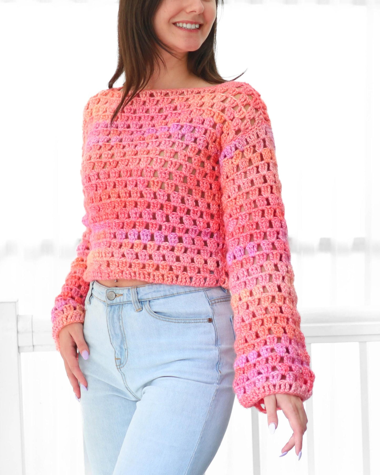 May include: A cropped, long-sleeved crochet sweater in shades of pink and orange. The open weave design reveals the skin beneath. The sweater has a wide neckline and is paired with light blue jeans.