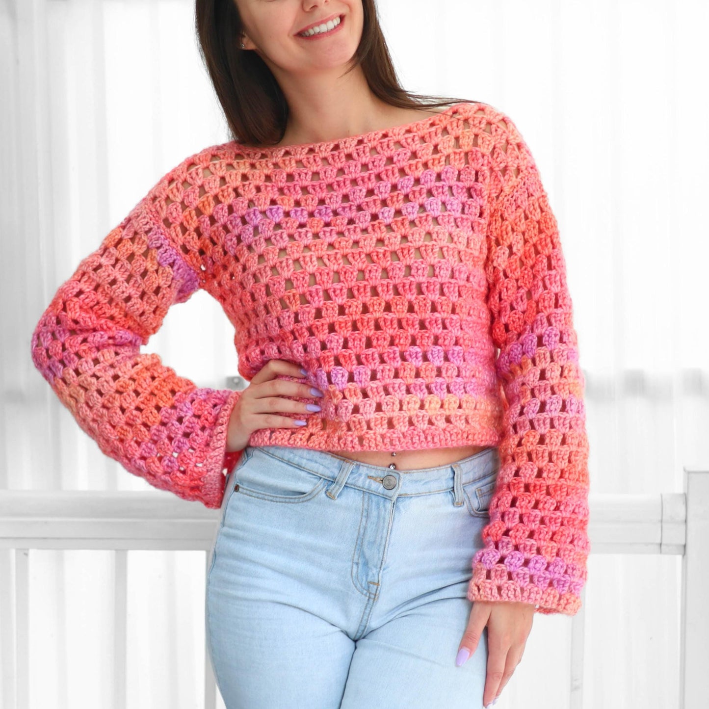 May include: A crocheted sweater in shades of pink, orange, and purple. The open weave design features long sleeves and a boat neck. The sweater is a cropped style, perfect for layering or wearing on its own.