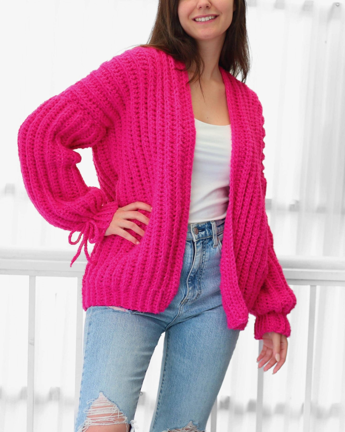 May include: A bright pink, chunky knit cardigan with long sleeves and a relaxed fit. The cardigan features a shawl collar and tie details at the cuffs. The model is wearing a white tank top and light blue jeans.