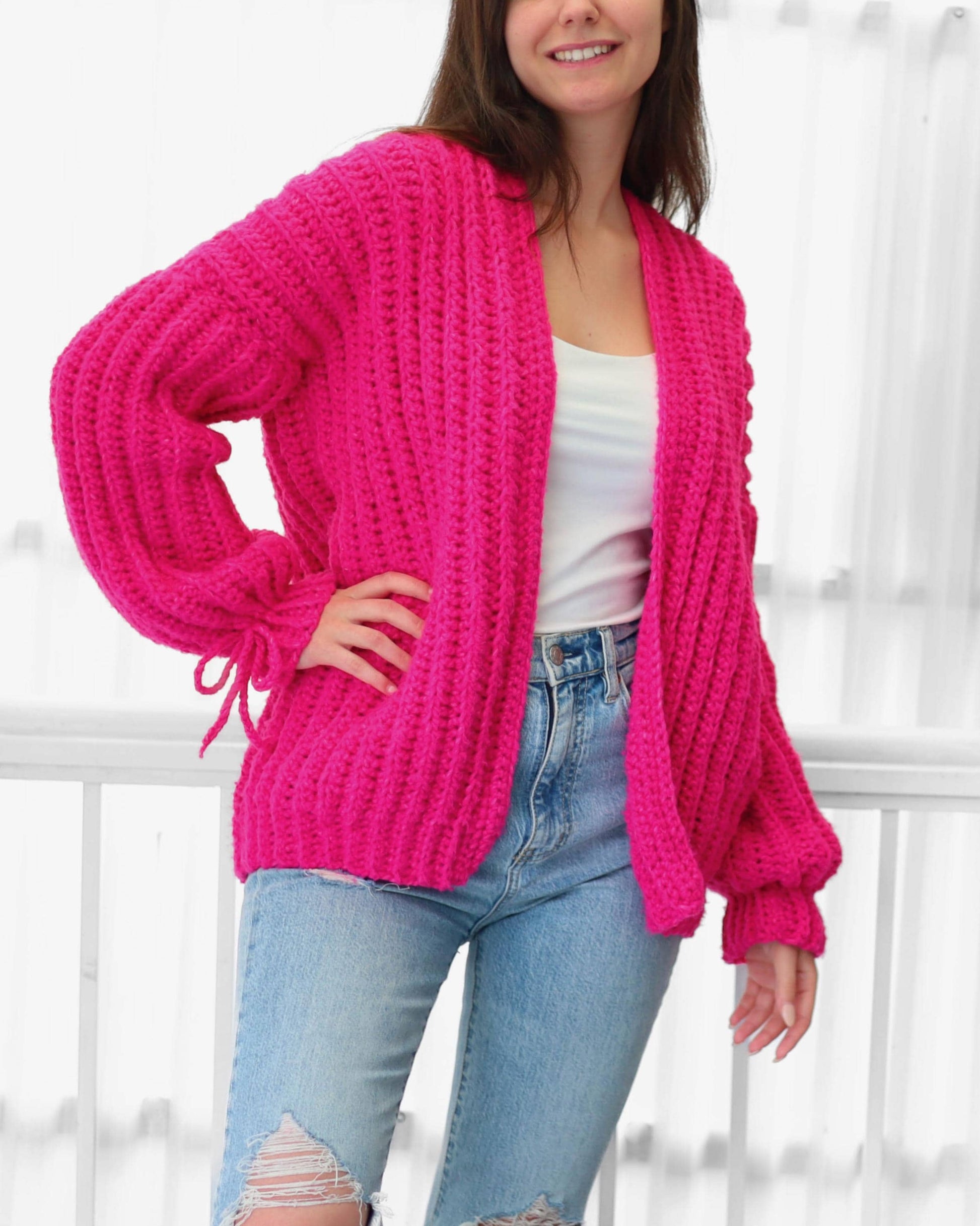 May include: A bright pink, chunky knit cardigan with long sleeves and a relaxed fit. The cardigan features a shawl collar and tie details at the cuffs. The model is wearing a white tank top and light blue jeans.