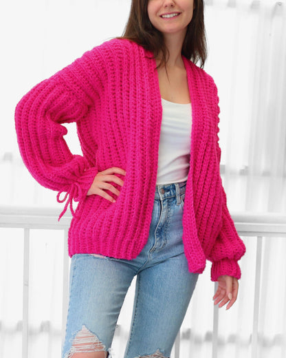May include: A bright pink, chunky knit cardigan with long sleeves and a relaxed fit. The cardigan features a shawl collar and tie details at the cuffs. The model is wearing a white tank top and light blue jeans.