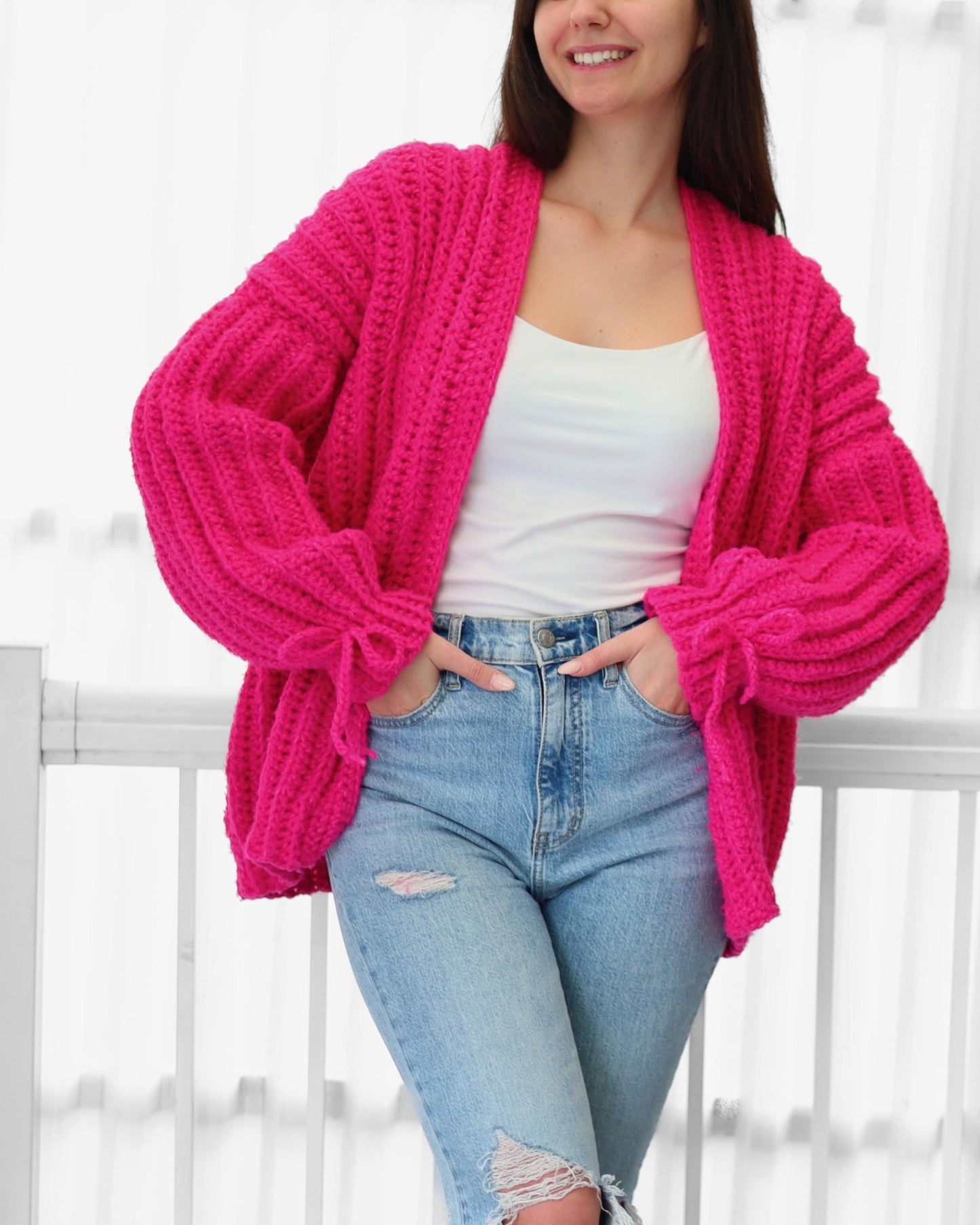 May include: A bright pink, chunky knit cardigan with long sleeves and a relaxed fit. The open-front sweater features a ribbed texture and is paired with light blue jeans. The sleeves have a tie detail at the wrist.