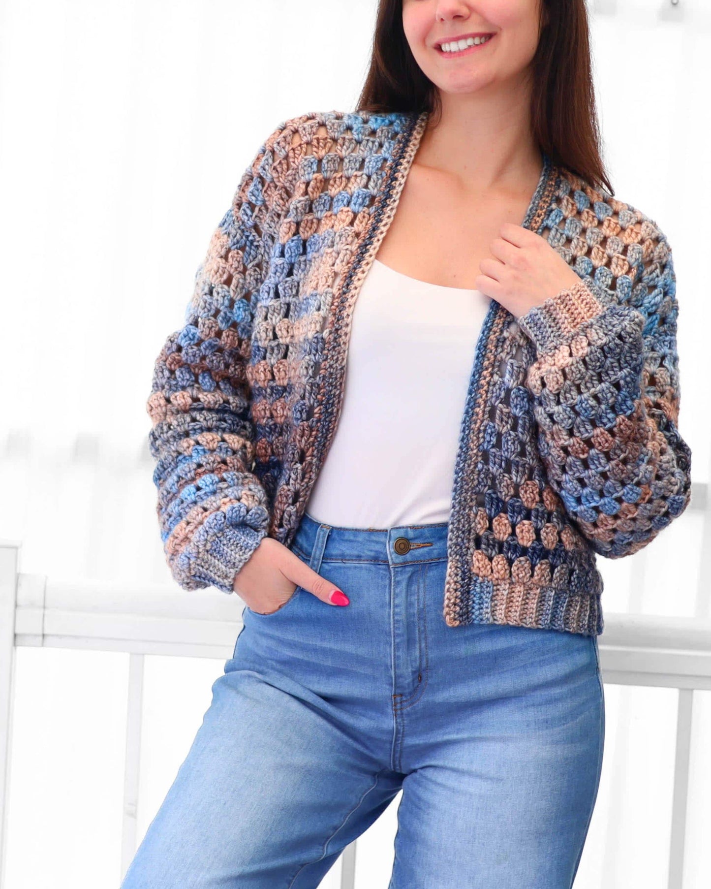 May include: A handmade crochet jacket in shades of blue, tan, and gray. The open-front cardigan features a textured stitch pattern and long sleeves. The jacket is paired with blue jeans.
