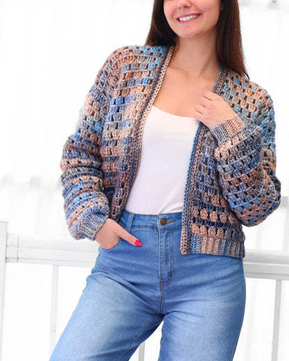 May include: A handmade crochet jacket in shades of blue, tan, and gray. The open-front cardigan features a textured stitch pattern and long sleeves. The jacket is paired with blue jeans.