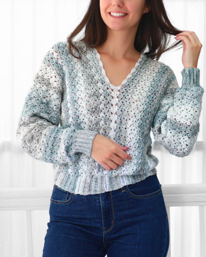 May include: A crochet sweater in shades of blue, white, and gray. The sweater has a V-neck and long sleeves with ribbed cuffs. The bottom hem is also ribbed. The sweater is paired with blue jeans.