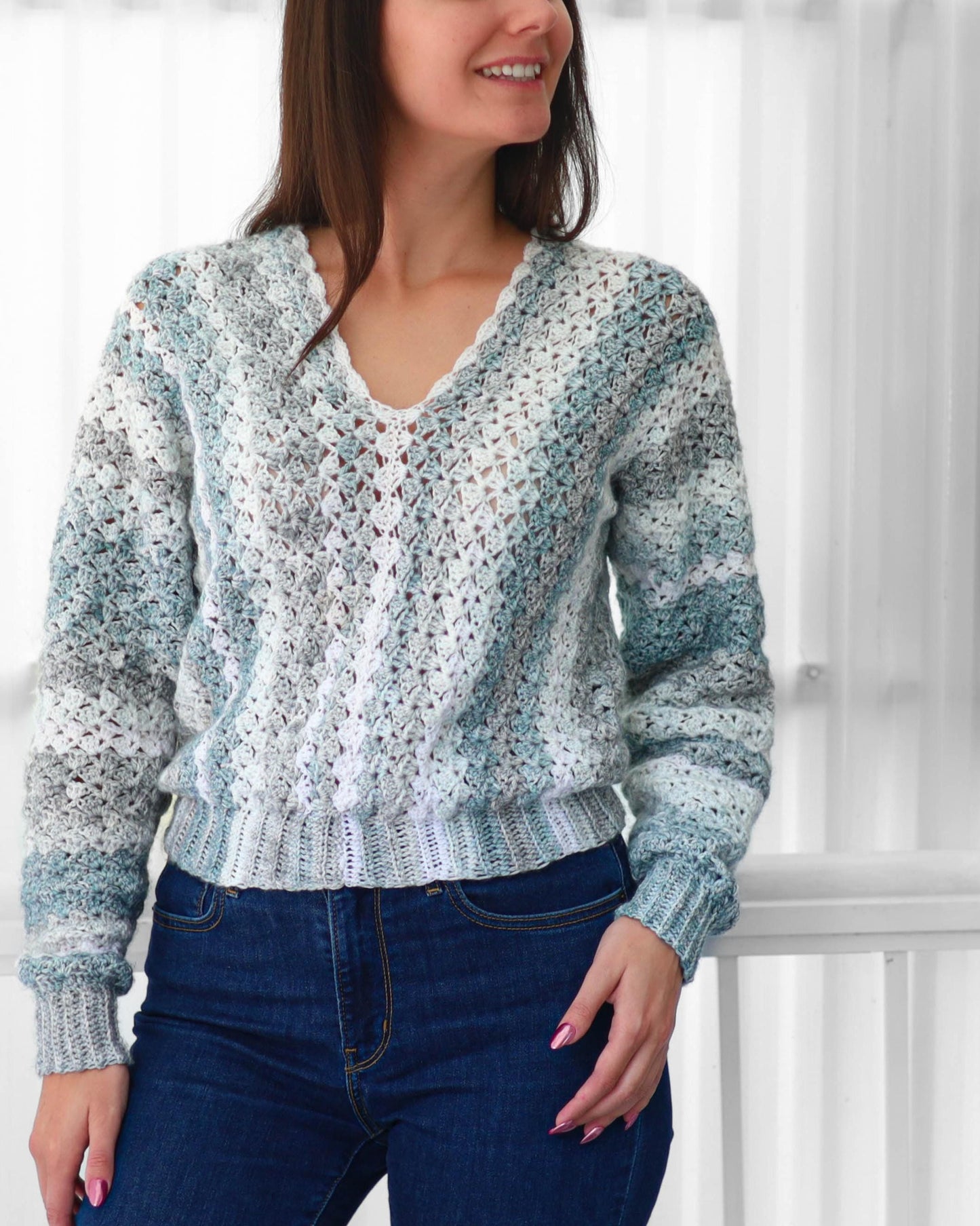 May include: A V-neck crochet sweater in shades of white, gray, and blue. The long-sleeved sweater has a ribbed hem and cuffs. The model is wearing blue jeans.